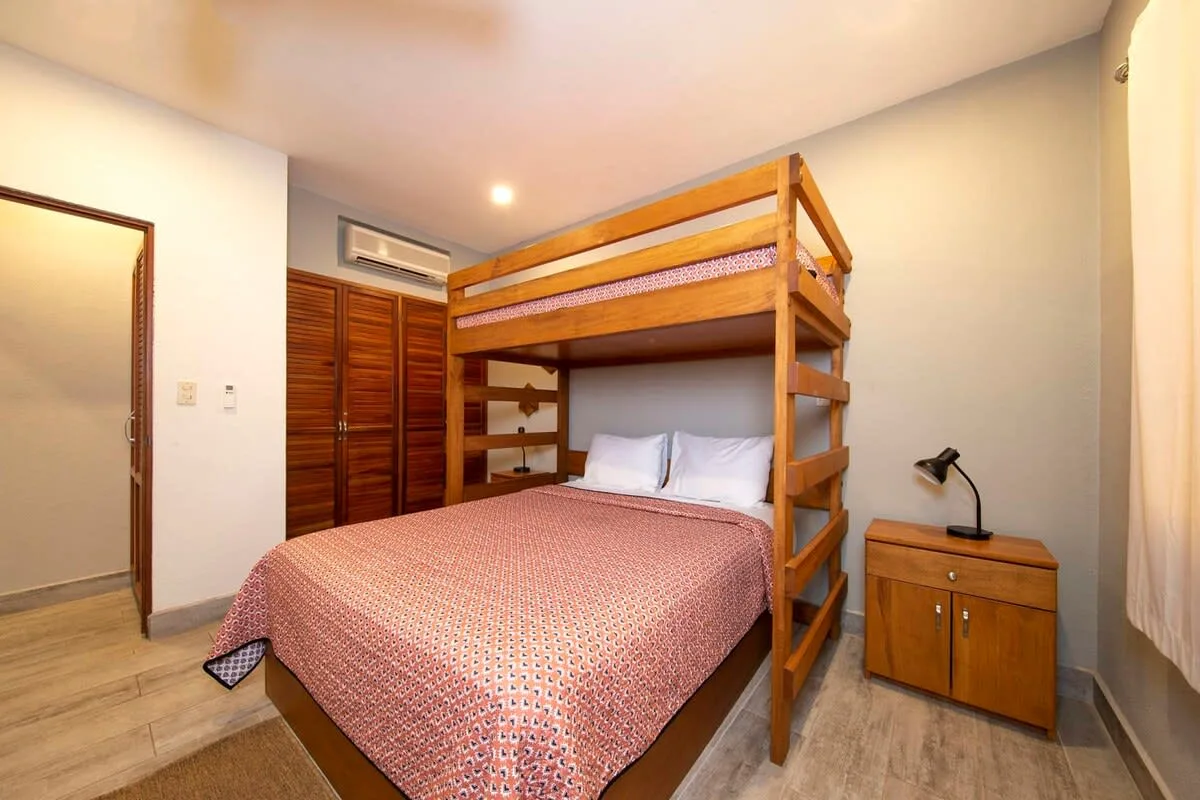 A bedroom with a wooden bunk bed above a larger bed and a bedside cabinet.