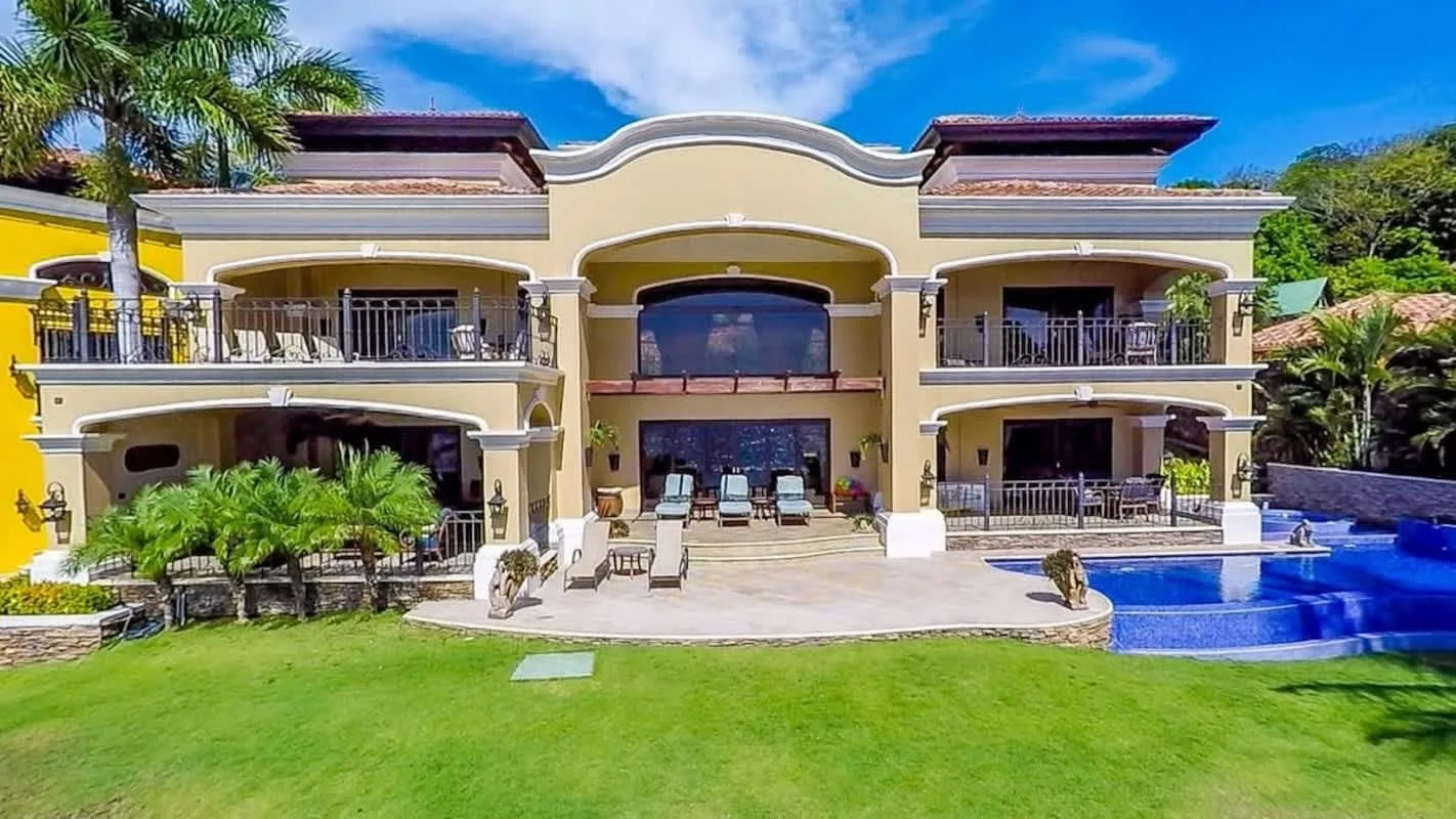 A large cream-colored villa stands behind a lawn, with a swimming pool beside the patio.