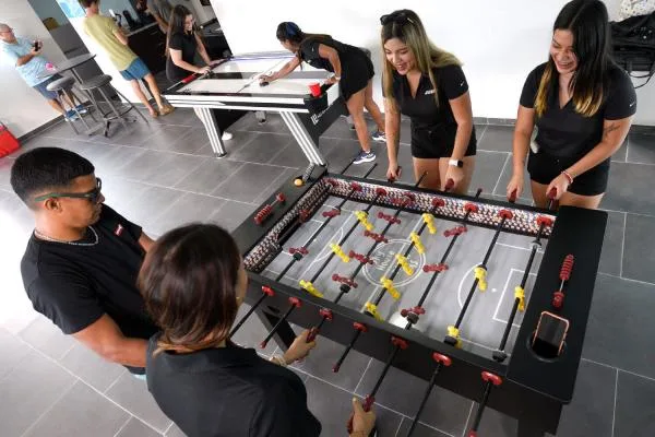 Varias personas juegan al futbolín alrededor de una mesa en una sala recreativa luminosa.