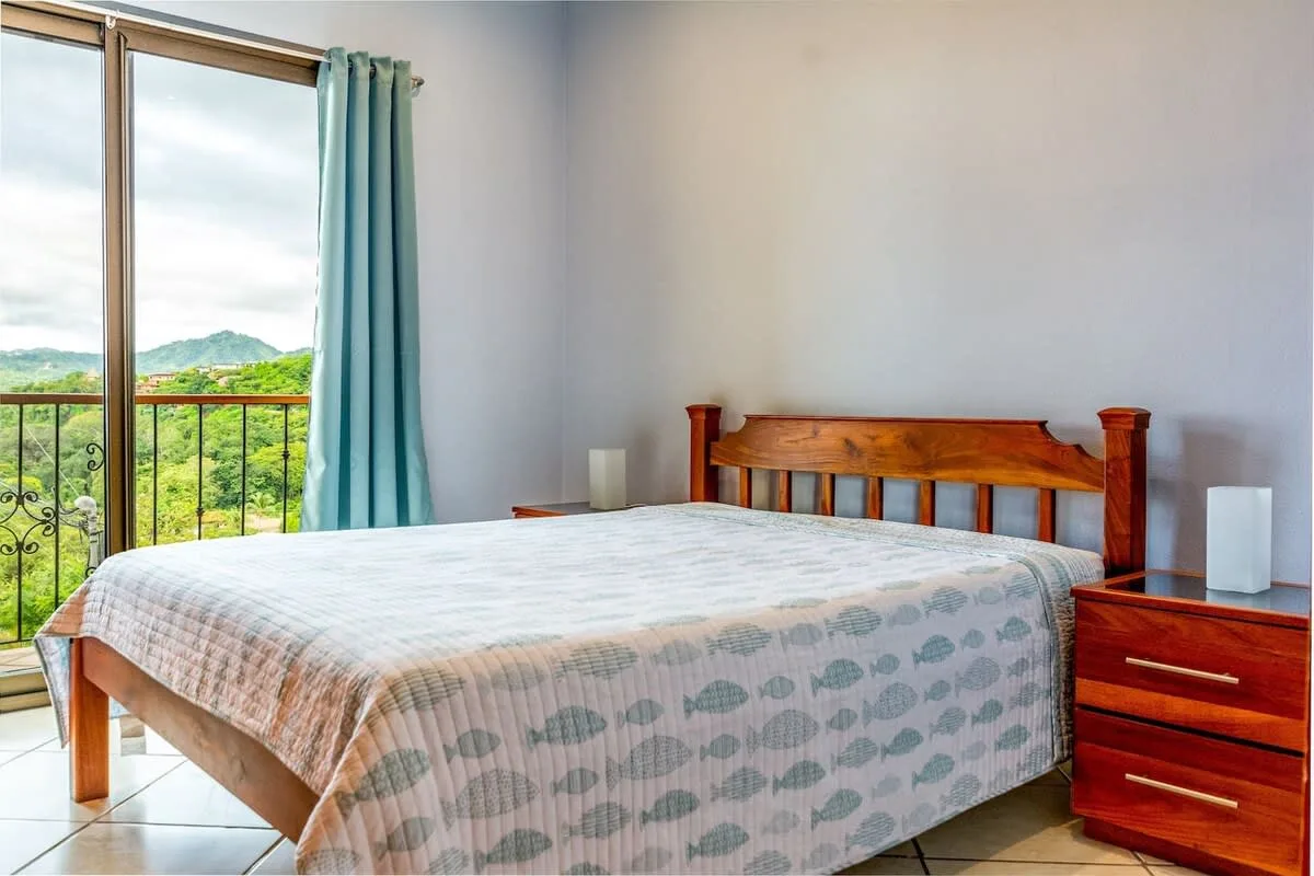 Dormitorio con una cama de madera junto a una ventana grande con vistas a la vegetación.