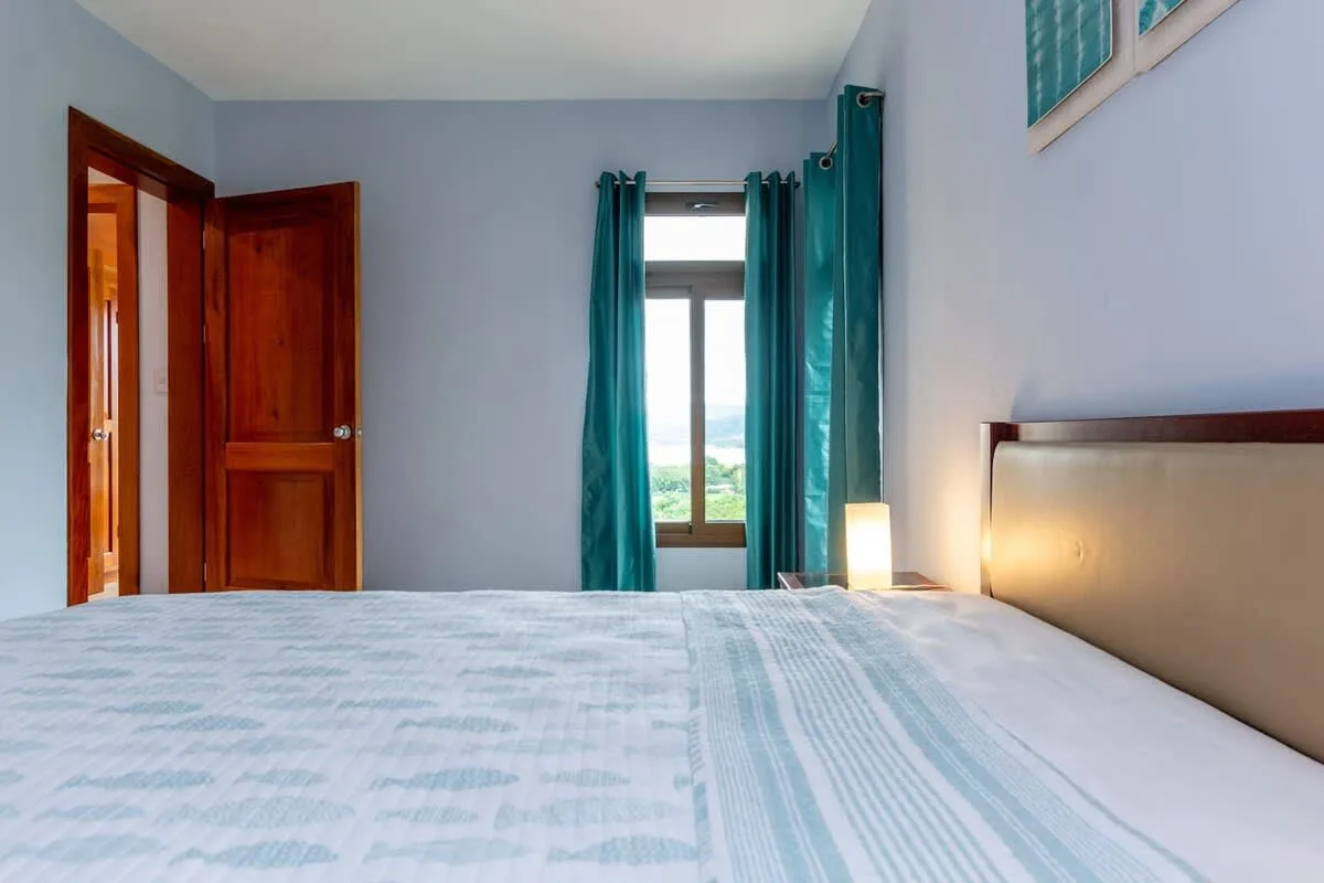 Un dormitorio con una cama, una puerta de madera abierta y cortinas turquesas junto a la ventana.