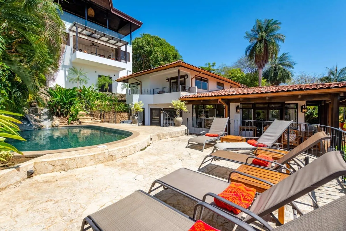 A sunlit poolside patio with lounge seating beside a tropical house.