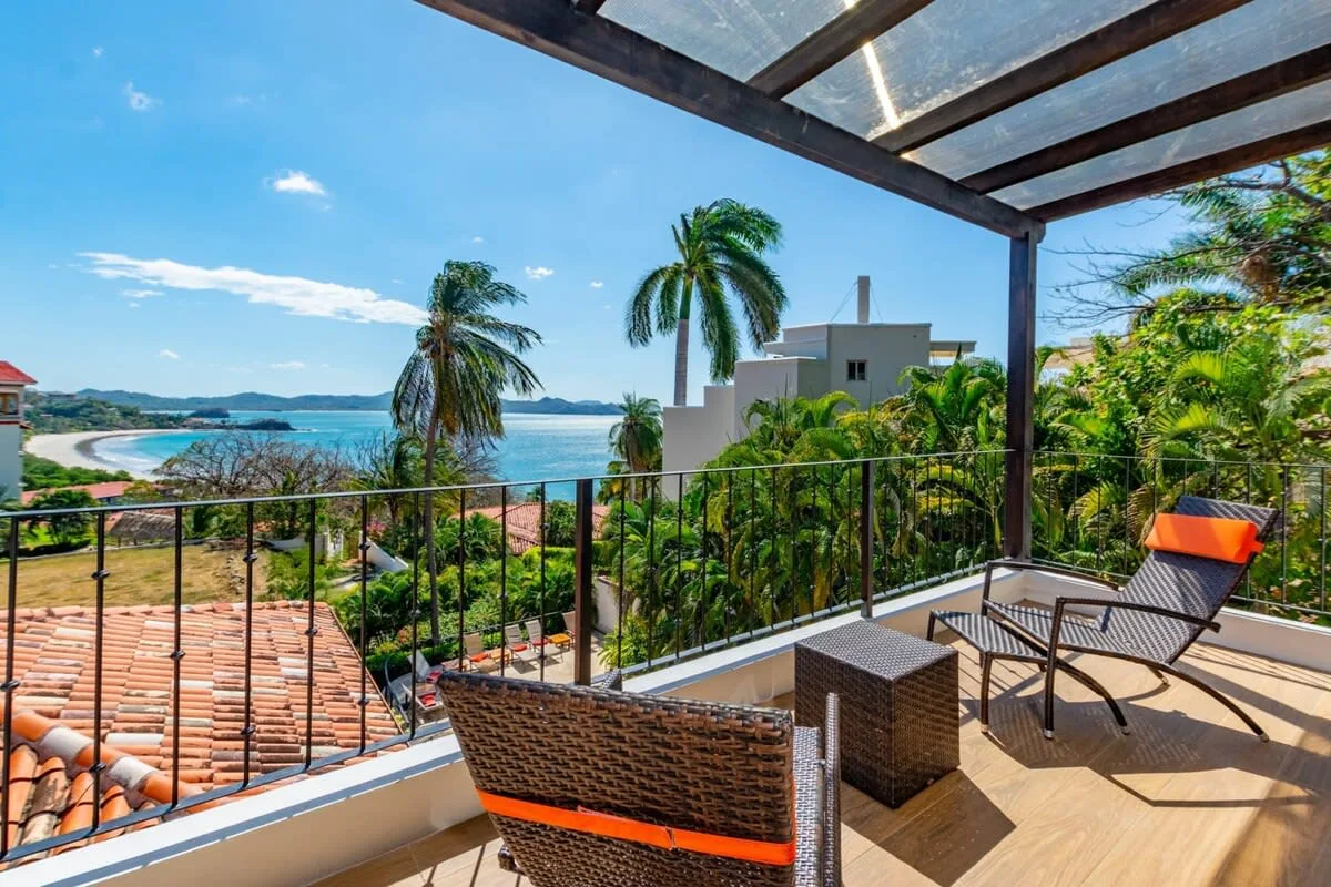 A covered balcony with seating overlooks palm trees and the ocean.