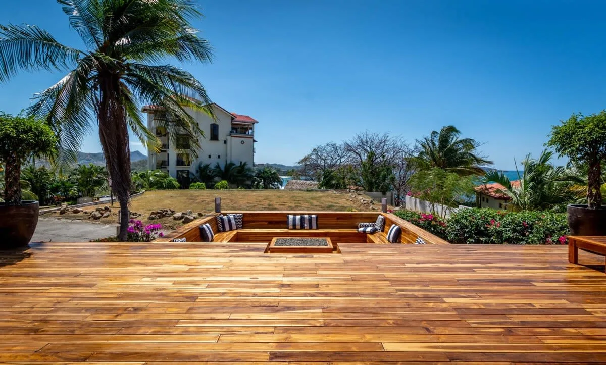 A sunny outdoor wooden deck overlooks a sunken seating area and palm trees.