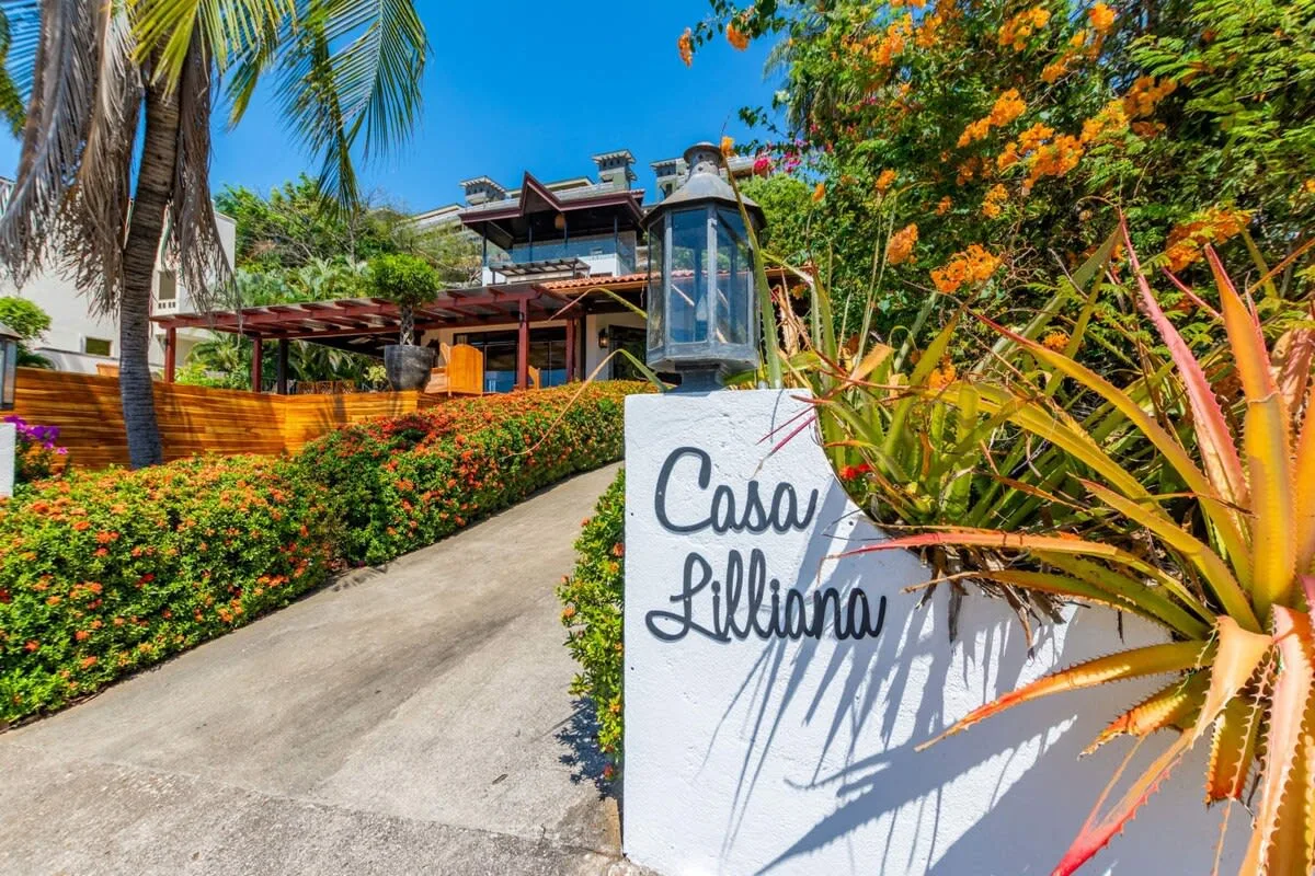 A tropical property entrance with a sloping driveway, flowering plants, and a sign.