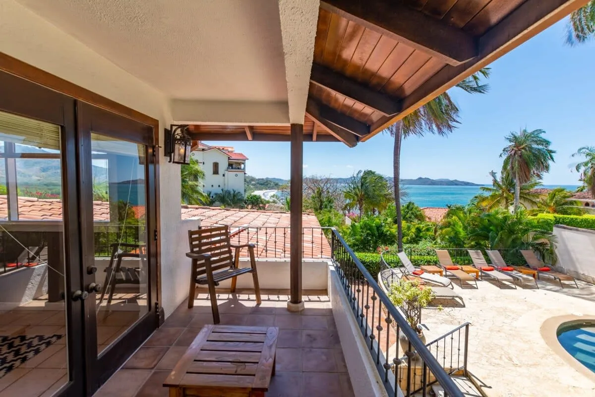 A covered terrace overlooks a pool, palm trees, and the sea.