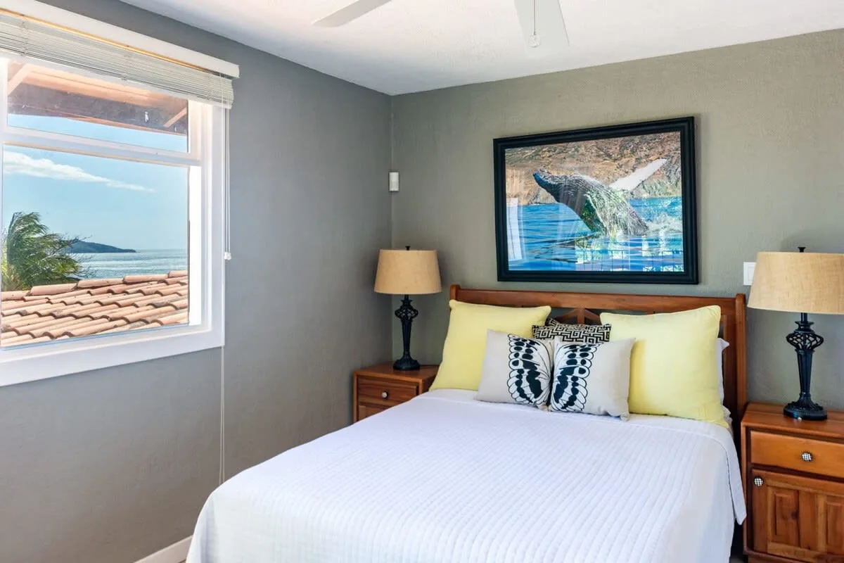 A bedroom with a bed beneath framed whale artwork and a window overlooking the ocean.
