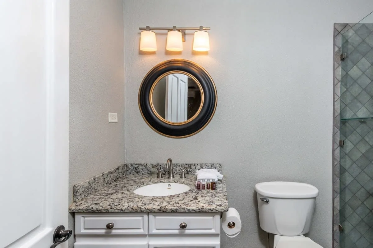 A bathroom vanity with a round mirror and toilet beside a glass shower.
