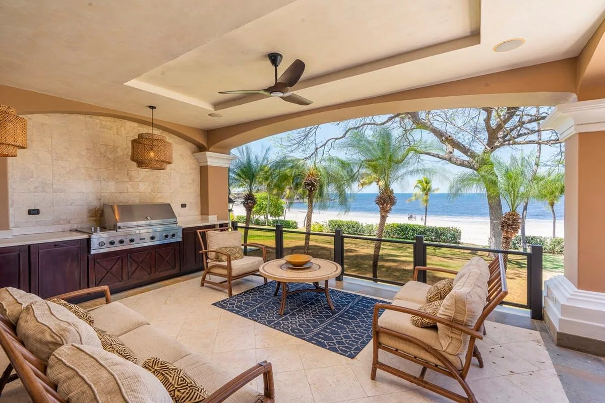 A covered patio with seating and a grill overlooks palm trees and the water.