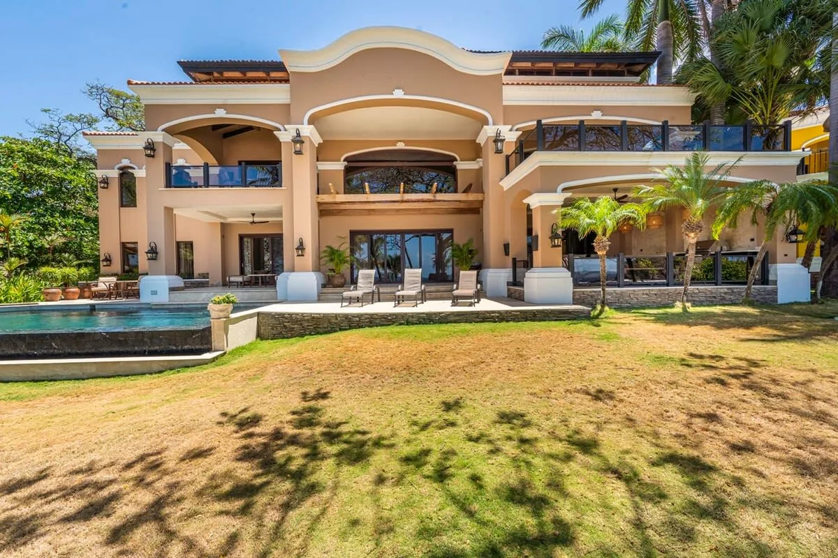 A large multi-story house faces a lawn, with a pool and outdoor seating.