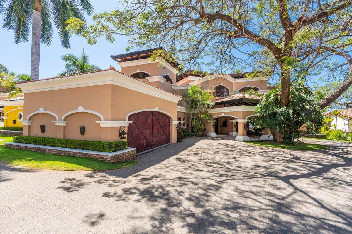 A large stucco house with a red gate and a tree-shaded driveway.