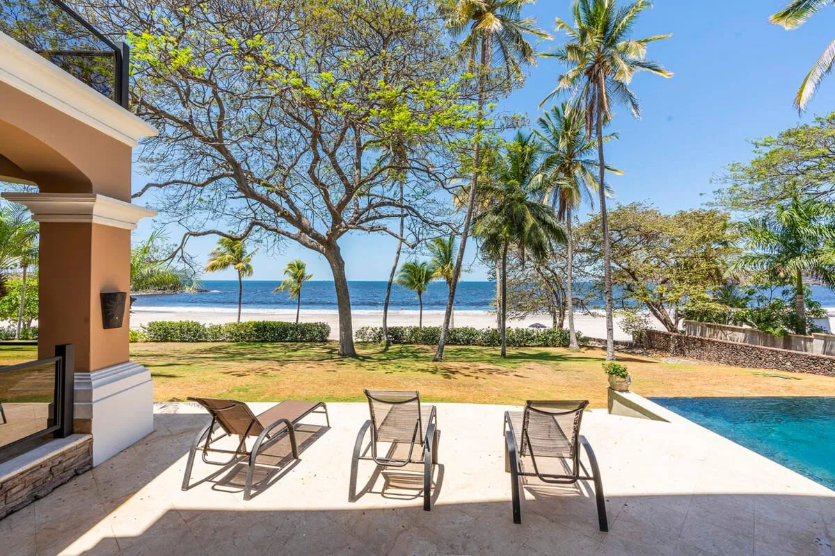 A sunlit patio with lounge chairs overlooks a beach and the ocean.
