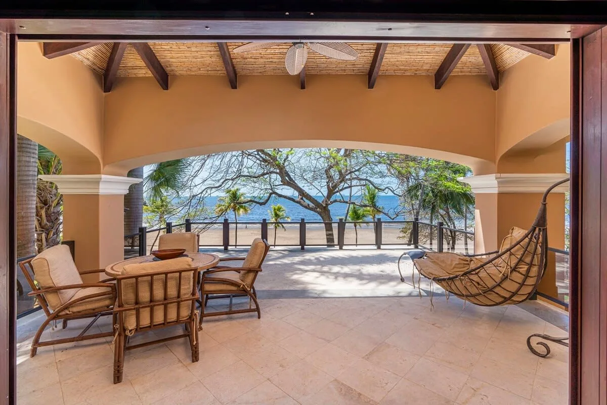 A covered patio opens onto a terrace with seating and a view of trees and water.