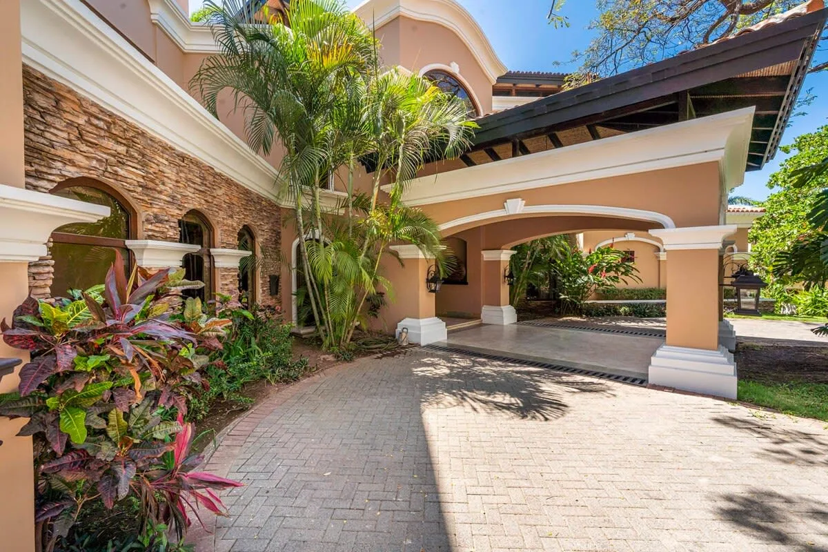 A covered driveway curves past a peach-colored building surrounded by tropical plants.