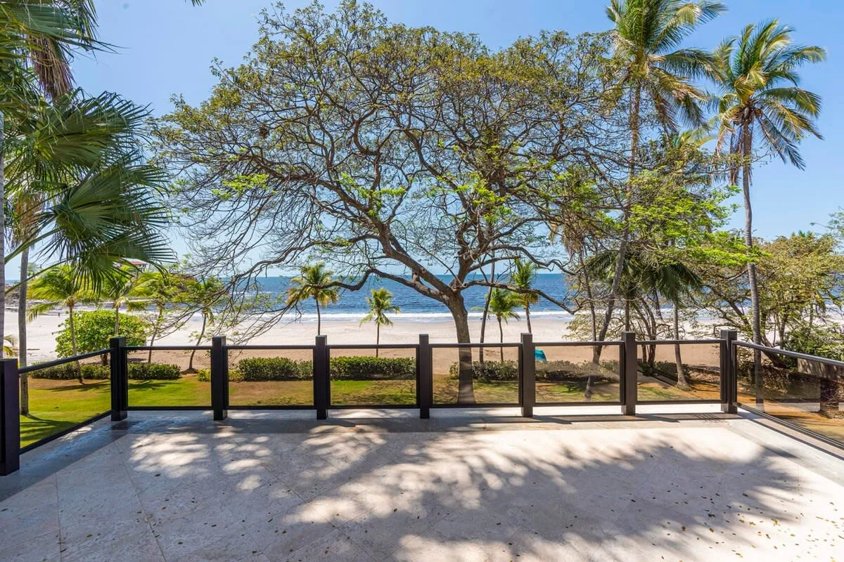 An outdoor patio overlooks a beach, with palm trees and a low railing.