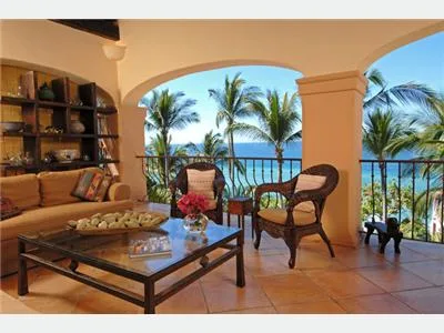 A covered terrace has a wicker seating area, a glass coffee table, and views of palm trees and the ocean.