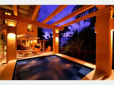 An illuminated patio overlooks a small swimming pool, with timber beams overhead and palm trees beyond.