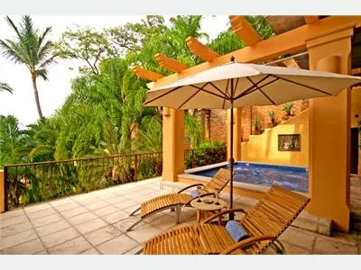 A tiled outdoor patio has wooden lounge chairs beneath a large umbrella, beside a small pool and tropical greenery.