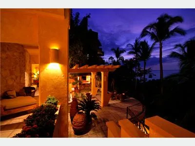 A warmly lit patio overlooks palm trees and the ocean beneath a deep blue evening sky.