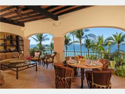 A covered terrace with wooden dining furniture overlooks palm trees and the ocean.