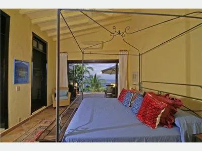 A canopied bed with colorful cushions sits in a room opening onto a tropical garden.