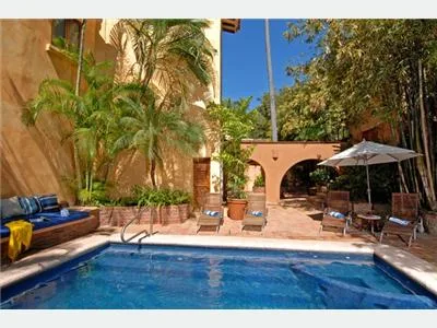 A blue swimming pool sits in a sunny courtyard with palm trees, patio chairs, and a shaded table.