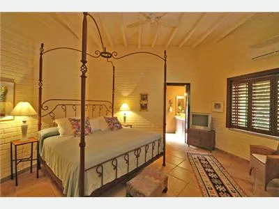 A warmly lit bedroom features a large wrought-iron four-poster bed, tiled floors, and a window with wooden shutters.