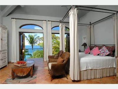 A bright bedroom has a canopy bed, wicker chairs, and open doors overlooking palm trees and the sea.
