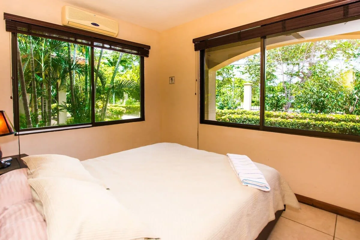 A bedroom with a bed beside two wide windows overlooking greenery.
