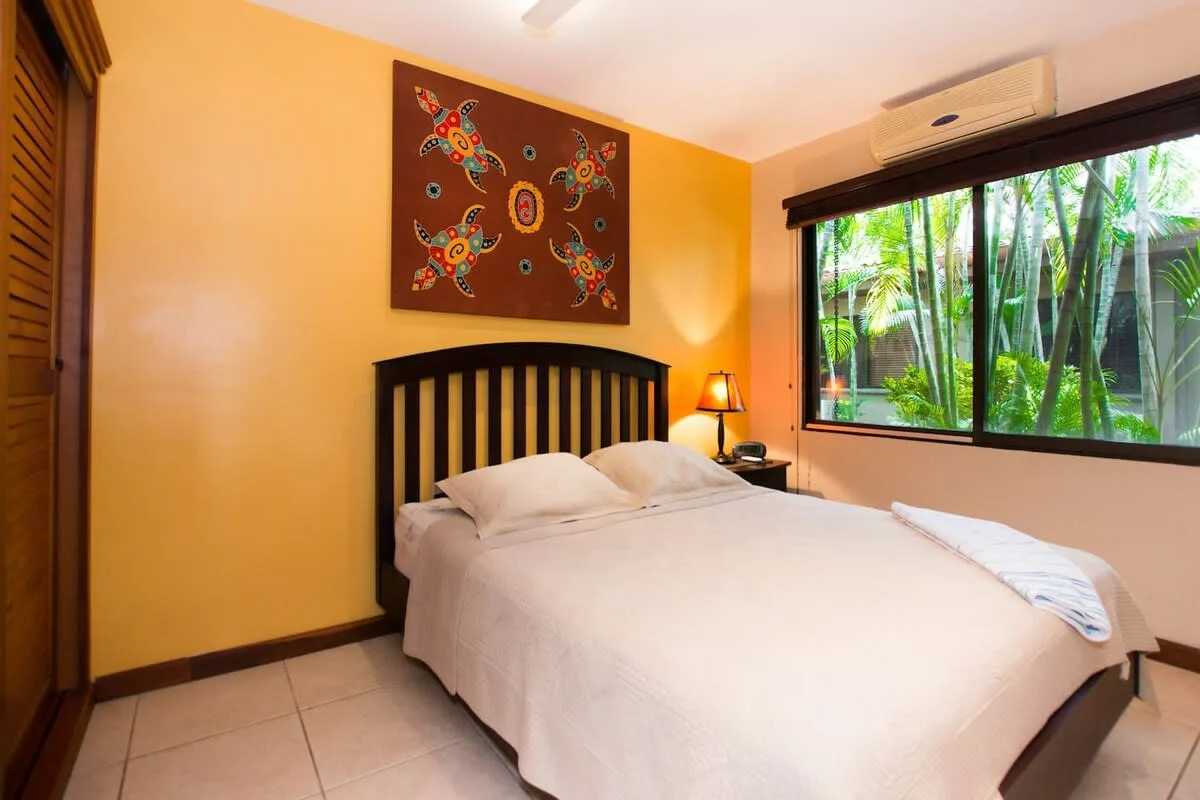 A bedroom with a neatly made bed and a large window overlooking greenery.