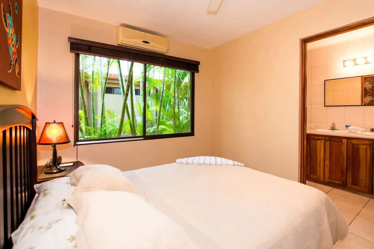 A bedroom with a bed beside a large window overlooking greenery.