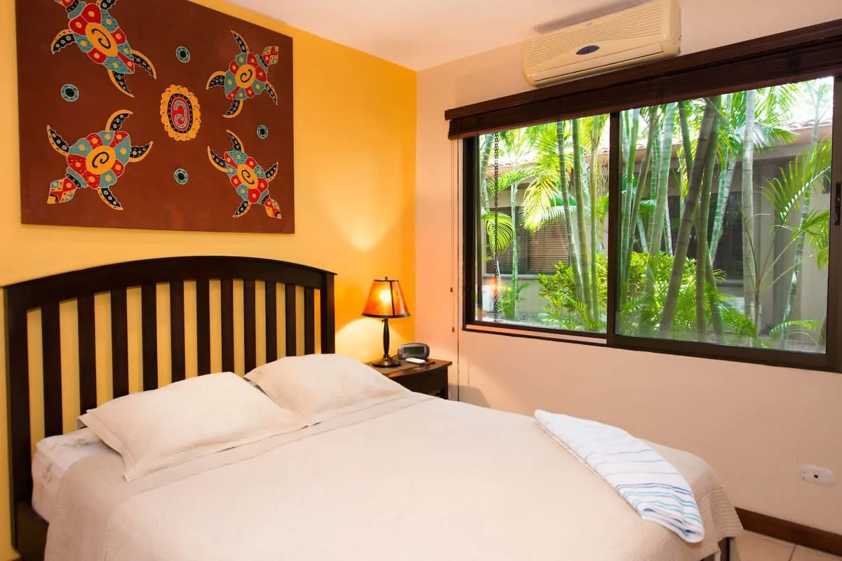 A bedroom with a neatly made bed beside a large window overlooking greenery.
