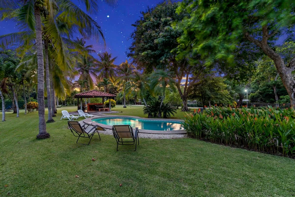 Una piscina de jardín está rodeada de palmeras, césped y plantas tropicales al anochecer.