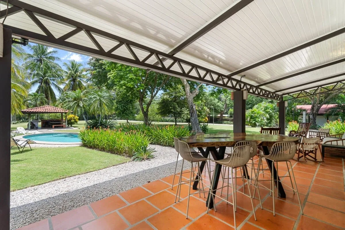 Un patio cubierto con asientos para comer da a un jardín y una piscina.