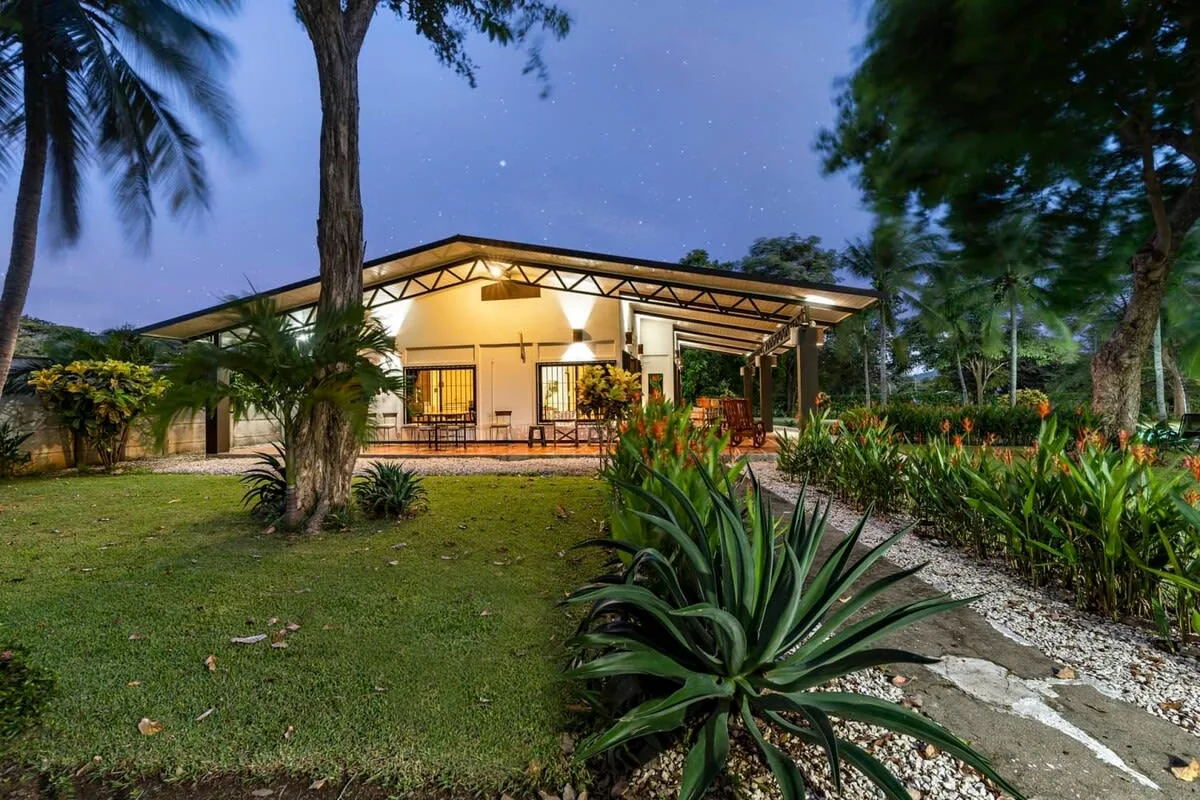 Un edificio iluminado con luz cálida se encuentra entre árboles y jardines al anochecer.