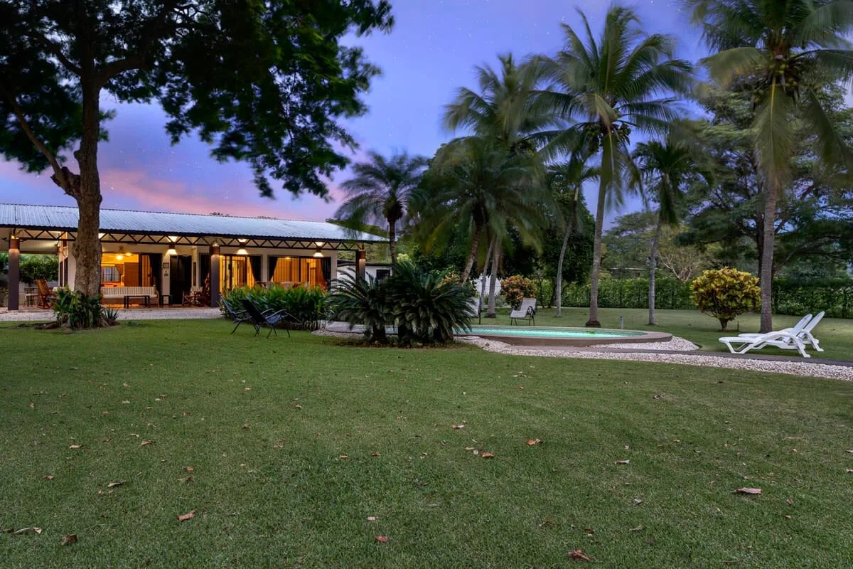 Un jardín con palmeras, una piscina y un edificio bajo al anochecer.