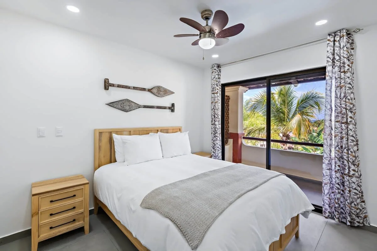 A bright bedroom with a neatly made bed and a large window overlooking palm trees.