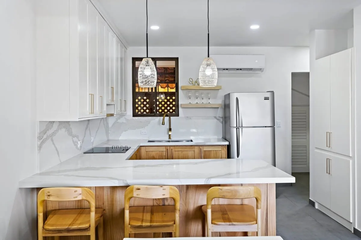 A bright modern kitchen with a white island, wooden seating, and pendant lights.