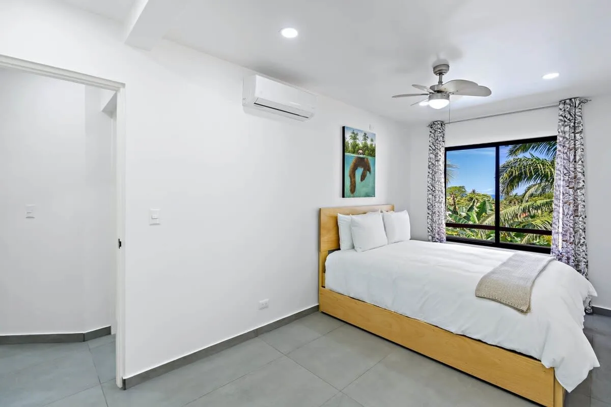 A bright bedroom with a bed beside a large window overlooking palm trees.