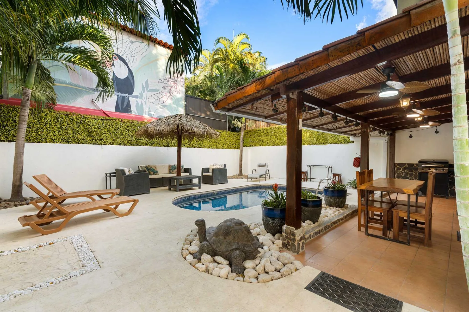 Una zona de piscina al aire libre junto a un comedor cubierto, con tumbonas y plantas tropicales.