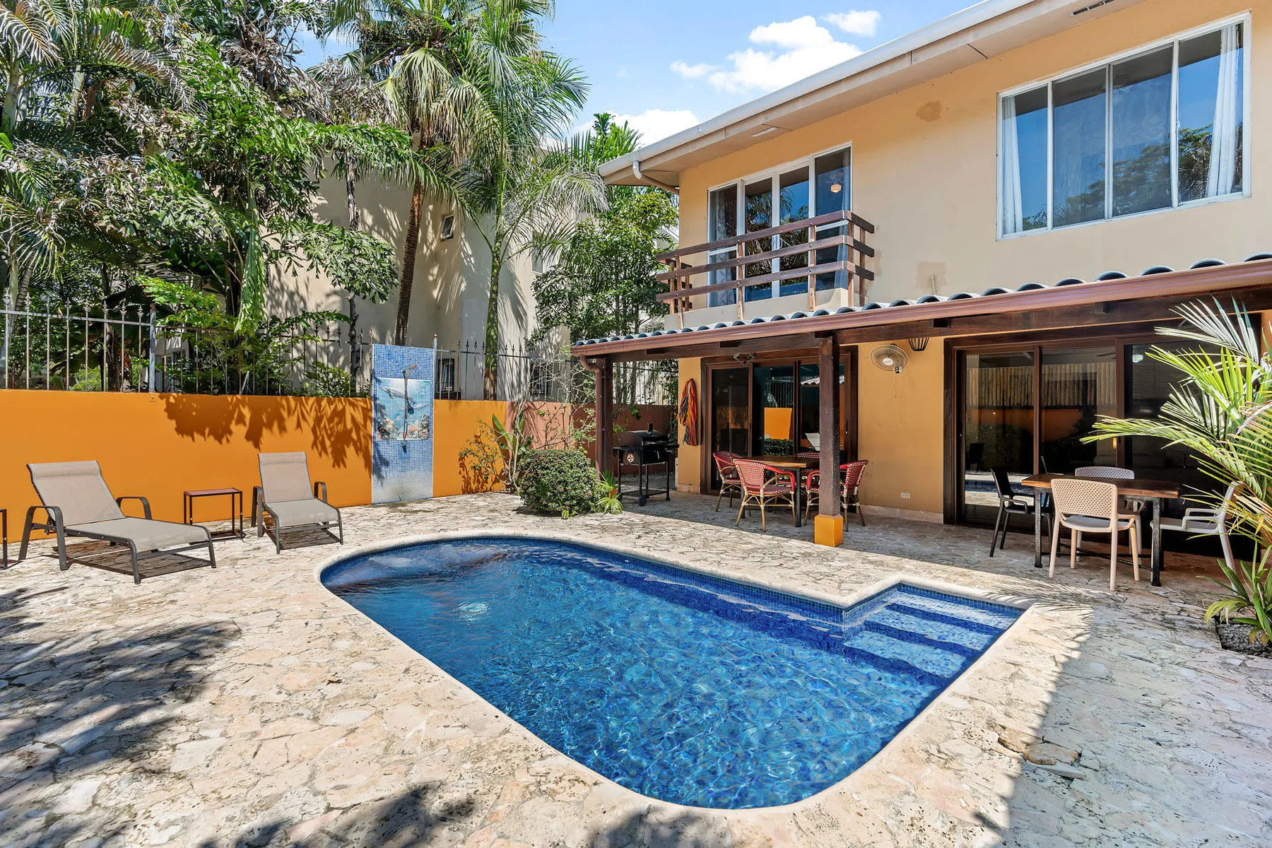 A swimming pool sits beside a patio with outdoor seating and a two-story building.