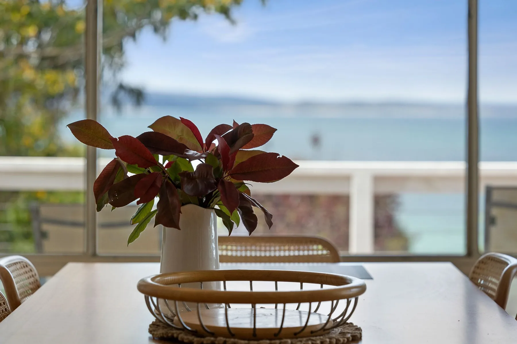 Una mesa de comedor con un arreglo de hojas está junto a una gran ventana con vista al agua.