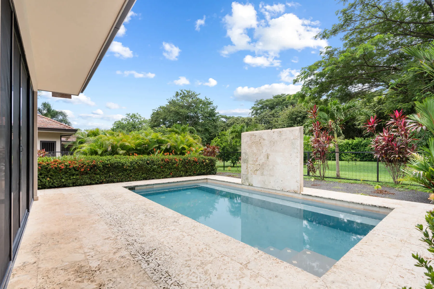 Una piscina al aire libre está junto a un patio pavimentado y un jardín frondoso.
