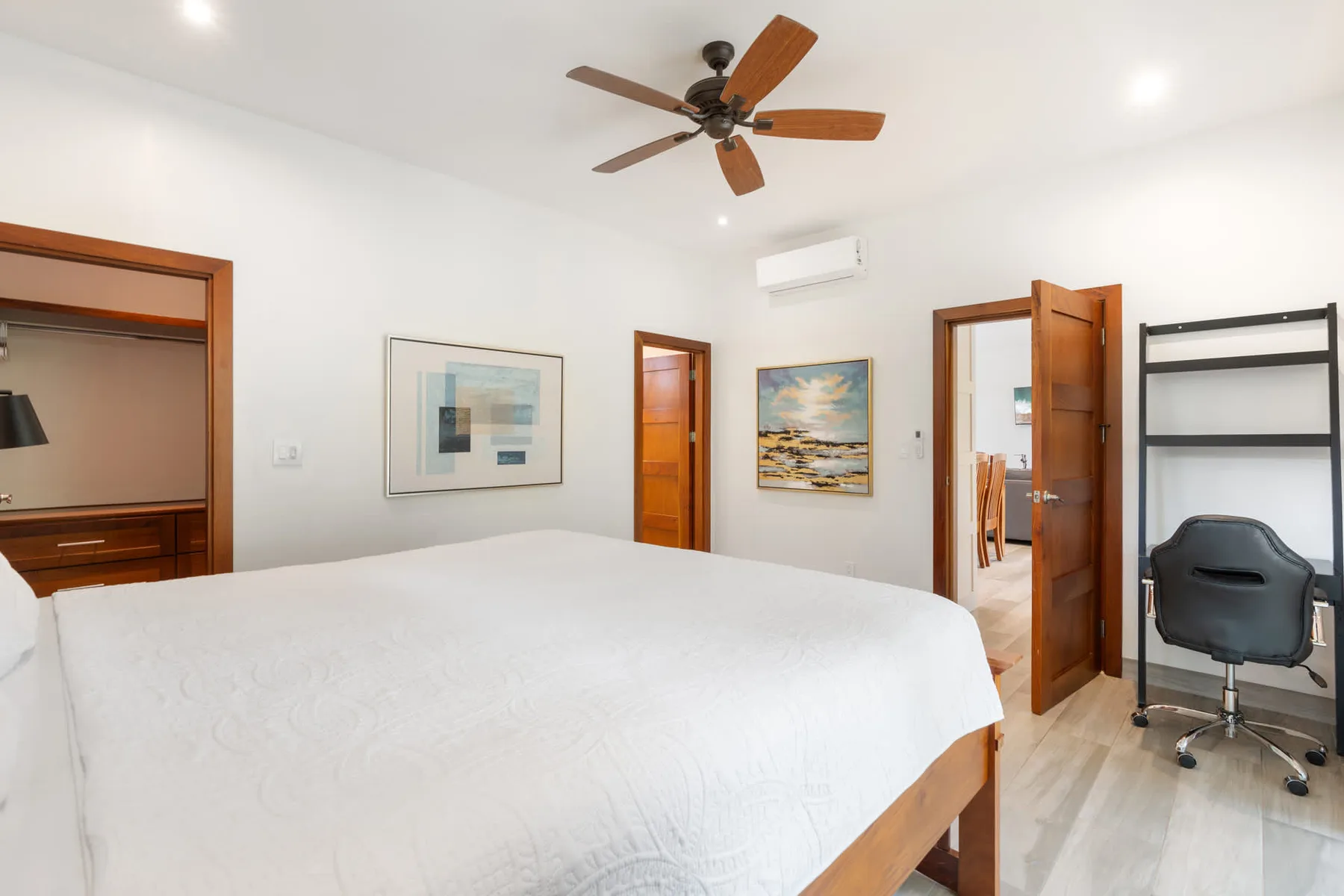 Un dormitorio luminoso con una cama grande, cuadros y una puerta de madera abierta.