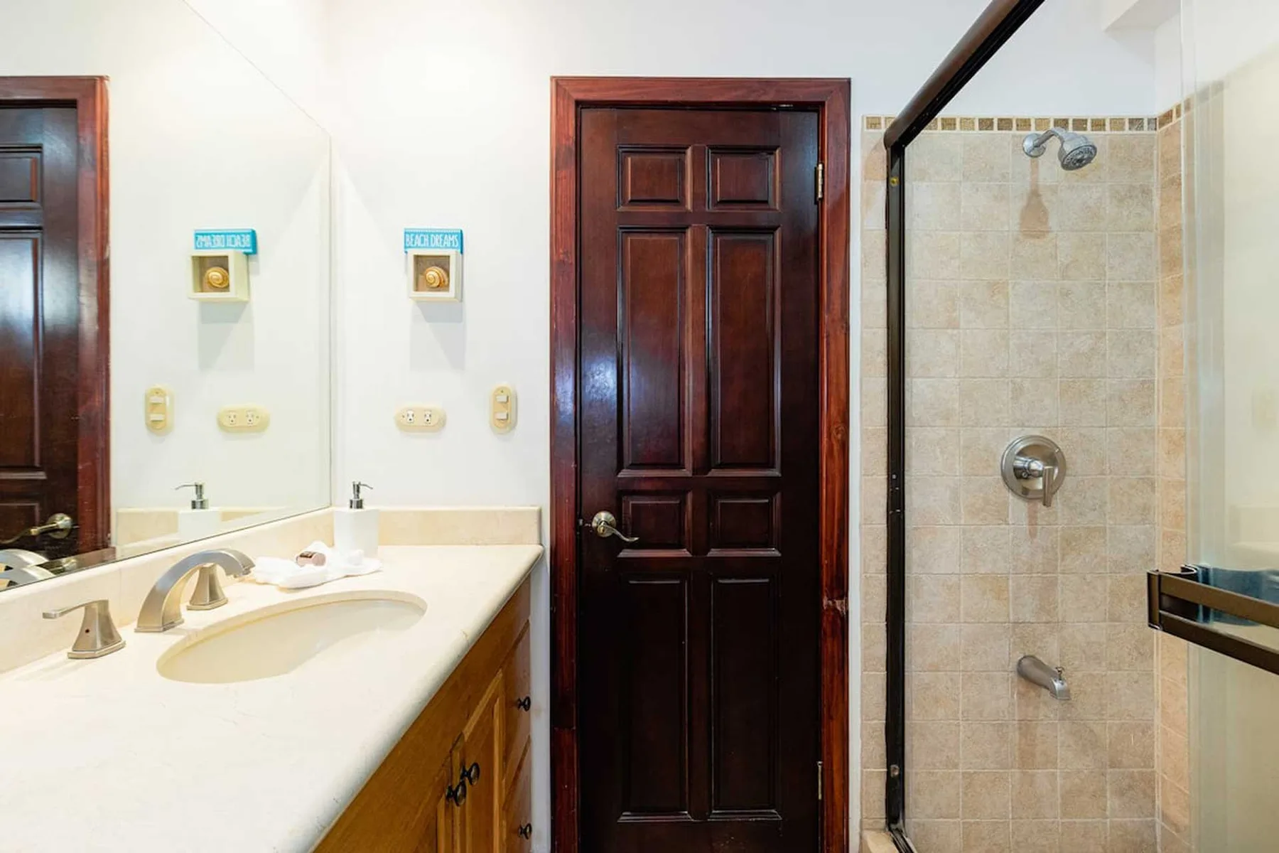 A bathroom with a vanity and a tiled shower beside a dark wooden door.