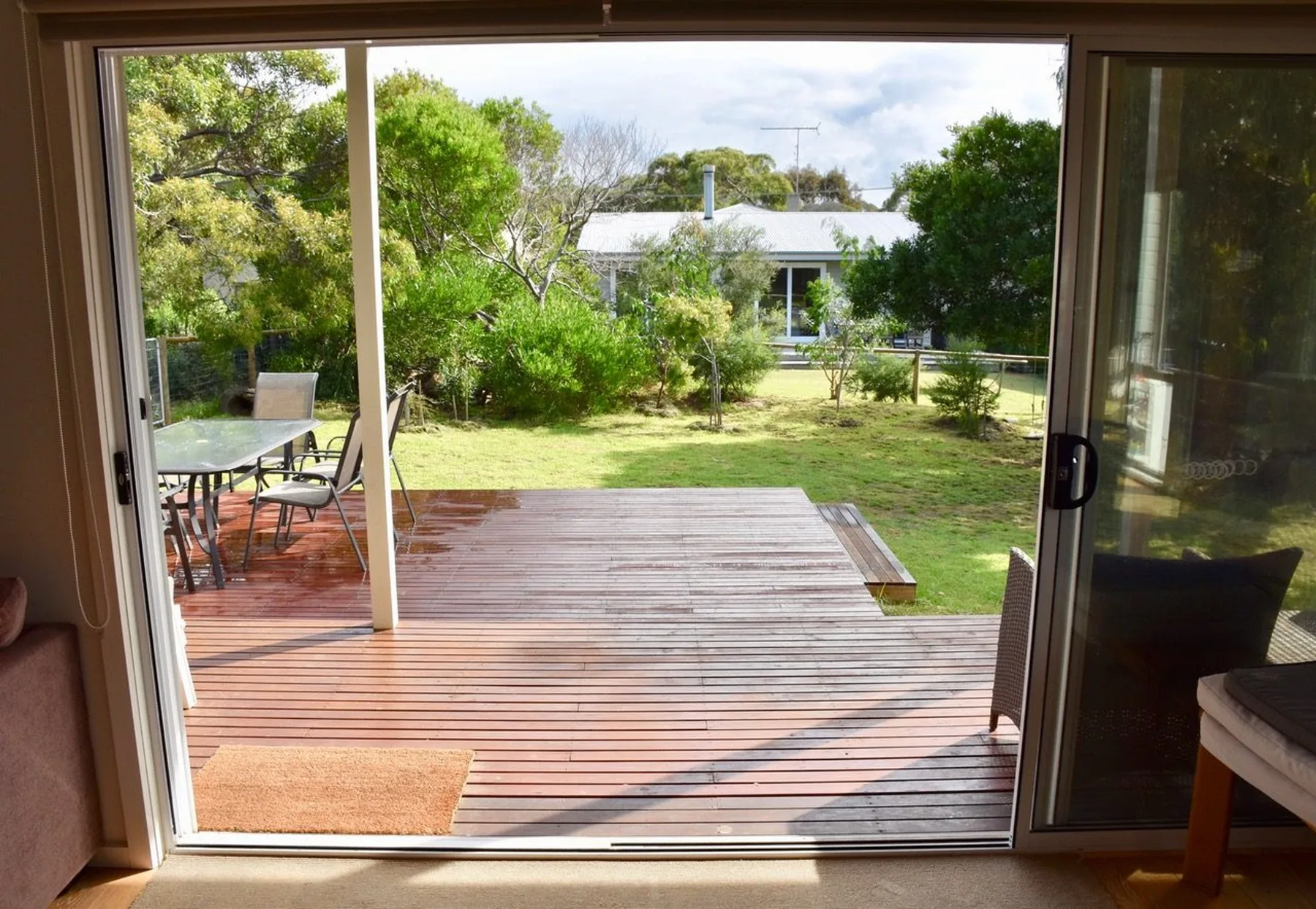 Una terraza de madera da a un jardín con césped, vista a través de puertas corredizas de vidrio.