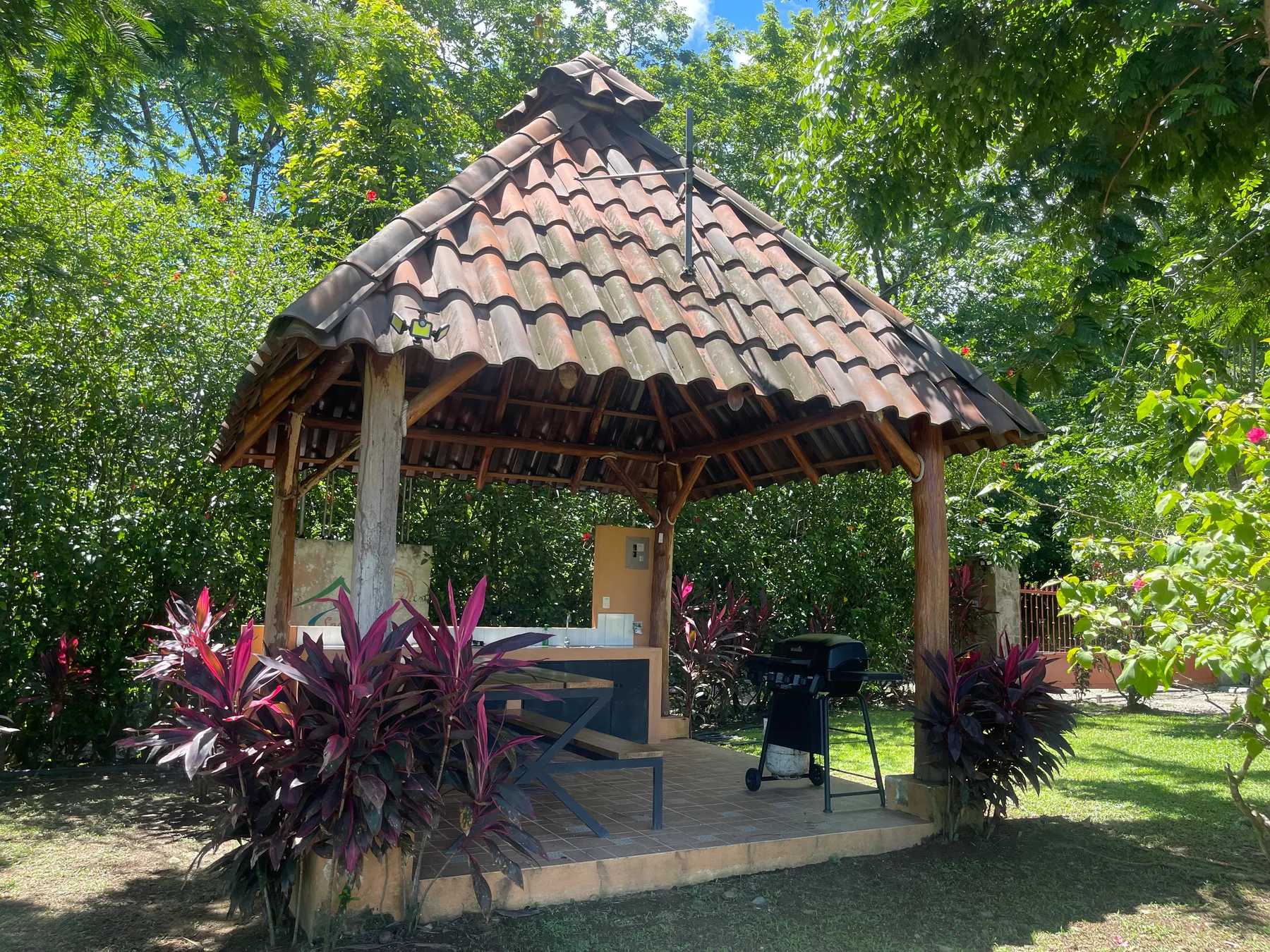 An open-sided garden pavilion with a tiled roof sits among trees and plants.