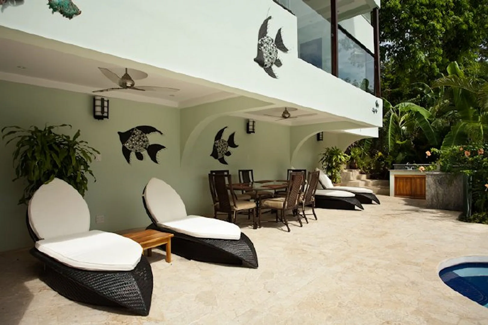 A poolside patio has lounge seating, a dining table, and tropical greenery.