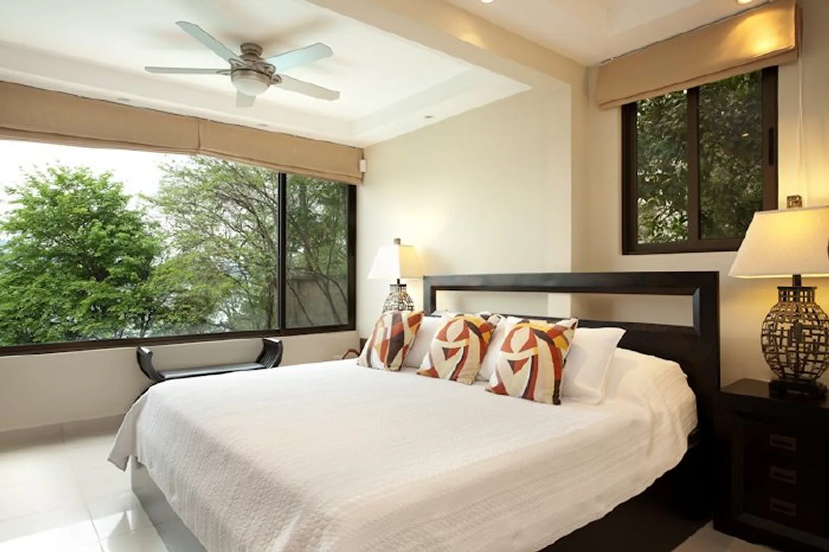 A bright bedroom with a large bed, bedside tables, and windows overlooking trees.