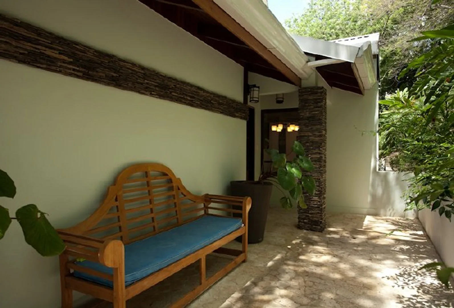 A shaded outdoor seating area has a wooden bench with a blue cushion.